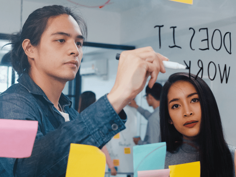 Design Thinking – Imageworks Two Asian professionals in a brainstorming session, penning their ideas on a clear board